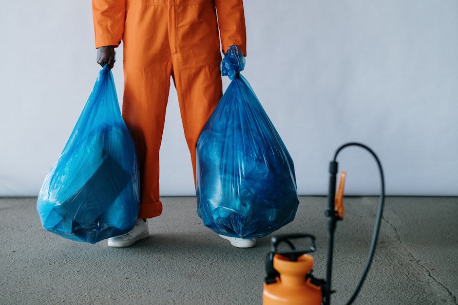 A person standing on a grey indoor floor, visible from the waist down, wearing orange overalls and white sneakers, holds a large, semi-transparent blue plastic rubbish bag in each hand. The bags appea