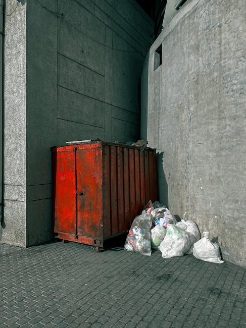 A waste collection worker dressed in a blue and orange high-visibility uniform is positioned beside a large white garbage truck, which is parked along a narrow urban street with cobblestone paving. Th