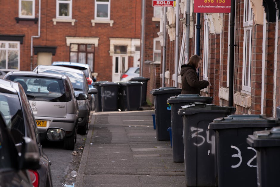 A photograph displays a cluttered scene of various discarded waste items layered on a driveway or pavement area. In the foreground, there are large, black plastic garbage bags filled with mixed rubbis