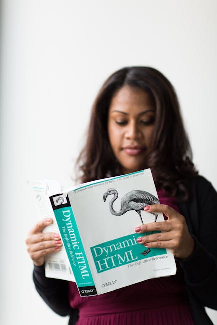 A woman with medium skin tone and dark brown, shoulder-length hair is standing indoors against a plain white wall, reading a book titled 'Dynamic HTML' by O'Reilly, featuring a black-and-white illustr
