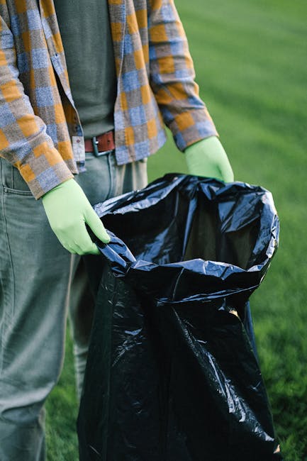 A person wearing bright blue disposable gloves is seen lifting a yellow plastic rubbish bag filled with various waste materials outdoors on a grassy surface. The bag appears to contain household waste